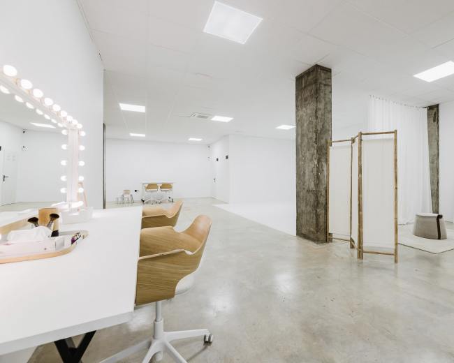 The image shows a spacious, minimalist room with white walls, concrete flooring, a vanity area with illuminated mirrors, and partitions separating different zones.