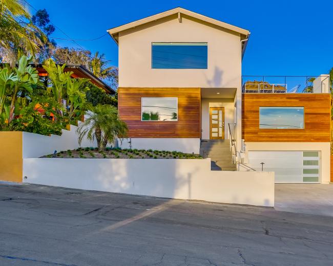 A modern two-story house with a mix of white and wooden exterior, large windows, and a landscaped front yard on a sloped street.