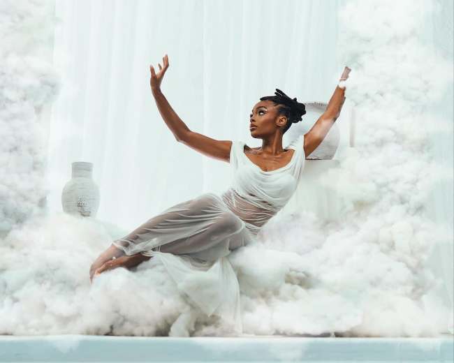 A dancer poses gracefully amidst a cloud-like backdrop with a reflective surface.
