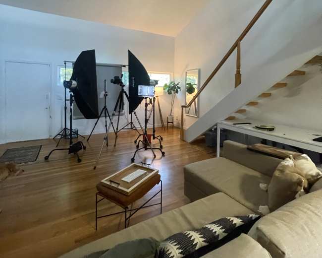 A brightly lit living room with video equipment set up, including tripods and softboxes, alongside a staircase and a sofa.