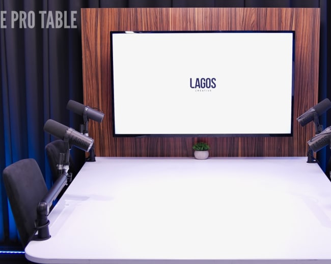 A modern podcast studio features a large screen displaying "LAGOS" on a wooden backdrop, with microphones positioned at two sides of a white table.