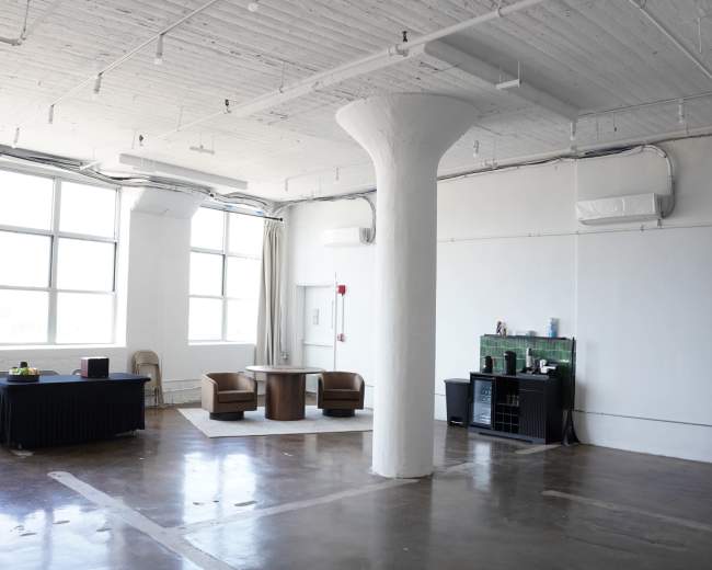 The image shows a spacious, well-lit room with large windows, a round table, two brown chairs, and a small kitchenette area against one wall.