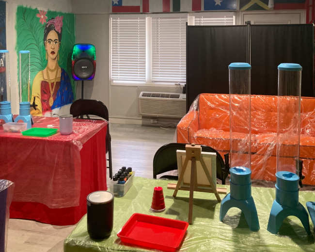 The room features colorful decorations, including a mural of Frida Kahlo, tables covered in plastic, and blue sky and cloud designs on the ceiling.