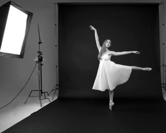 A dancer in a white dress performs an arabesque on a black stage surrounded by studio lighting equipment.