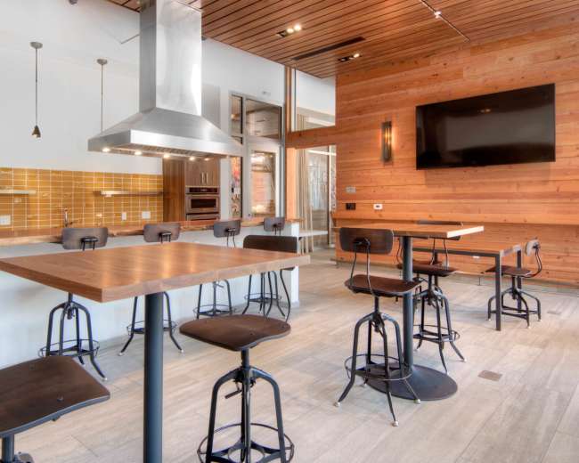 A modern kitchen with stainless steel appliances, wooden walls, and high-top tables with black stools.