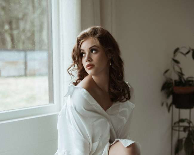 A young woman sits on a white stool by a window, wearing a loose white shirt with her legs crossed.