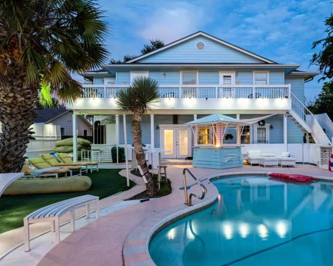The image shows a two-story light blue house with a swimming pool and lounge area in the foreground, surrounded by palm trees and outdoor furniture.