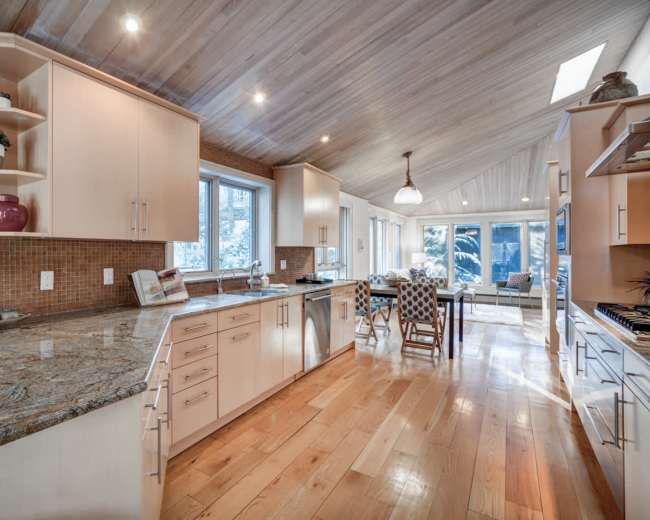 The image shows a modern kitchen with wooden floors, sleek cabinetry, and a large island, leading to a bright dining area with large windows.