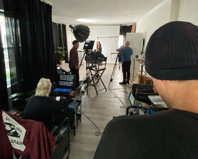 A small production team sets up equipment in a studio for a video shoot, featuring a woman in front of a backdrop and a teleprompter.