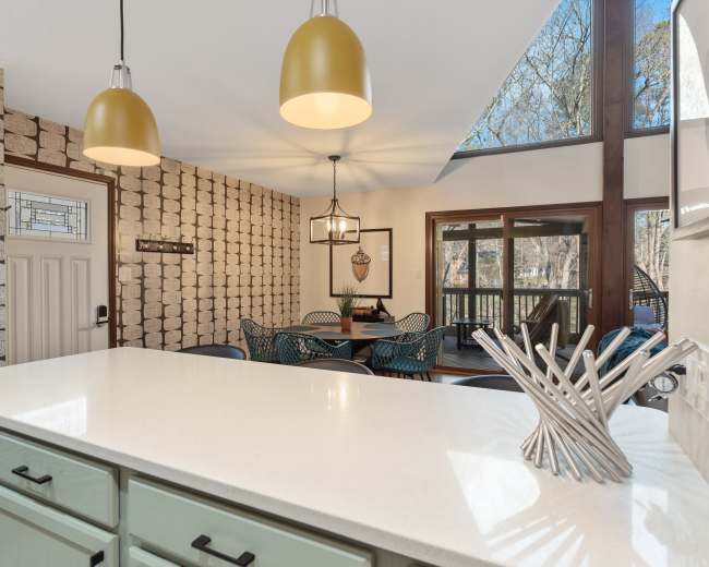 A kitchen area features a white countertop, green cabinets, and pendant lights, with a dining space visible through large windows that overlook an outdoor deck.