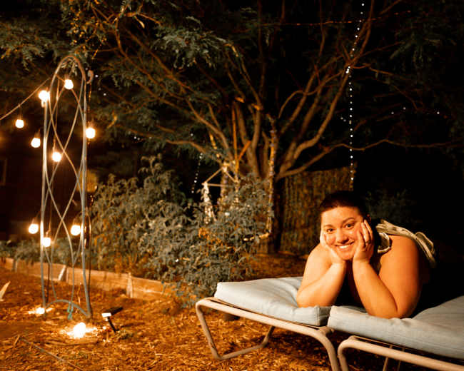 A person lies on a lounge chair in a dimly lit outdoor setting surrounded by trees and decorative string lights.
