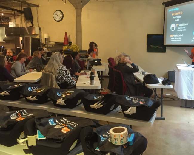 A speaker presents in front of an audience seated at long tables in a brightly lit industrial-style venue.