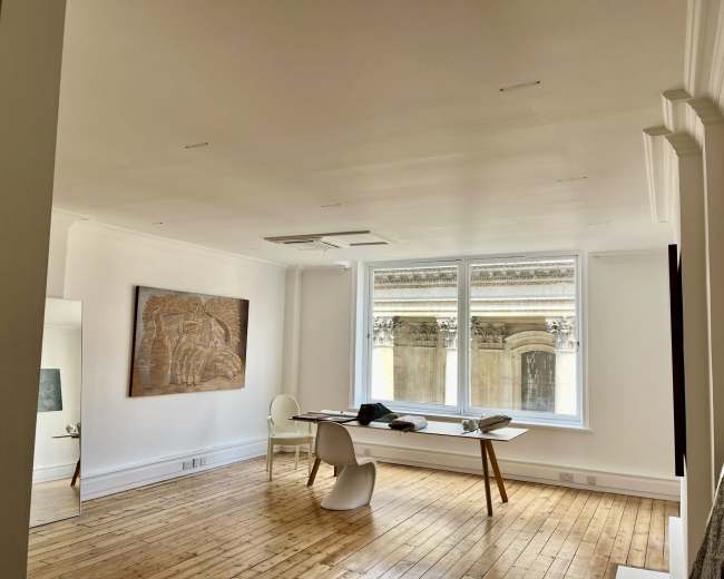 A modern, minimalist room featuring a wooden floor, a large window overlooking a building facade, a table with a few items on it, and a wall with an artistic panel.