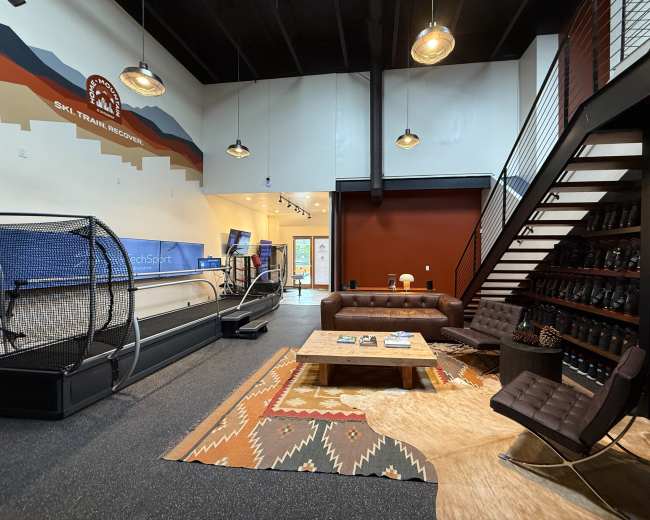 The image shows a spacious interior of a ski resort training facility featuring a treadmill, seating area with brown leather sofas, and a staircase leading to an upper level.