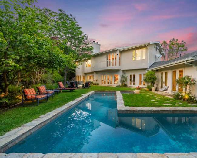 A spacious backyard features a swimming pool surrounded by lush greenery and modern lounge chairs near a two-story house at sunset.