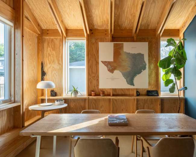The image shows a wooden room with a large map of Texas on the wall, a dining table, chairs, a modern lamp, and plants near the windows.