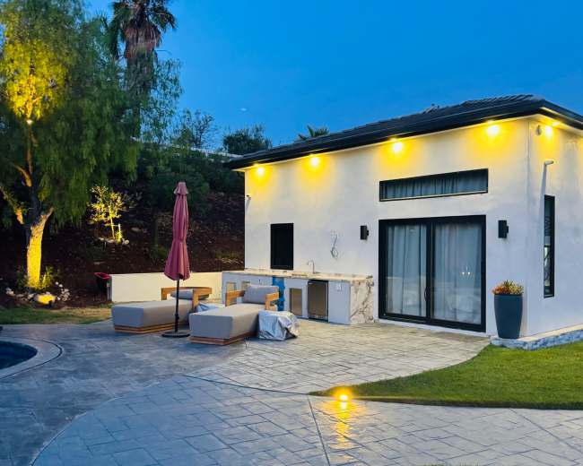 A well-lit outdoor patio features lounge furniture, an umbrella, and an outdoor kitchen beside a swimming pool, with palm trees in the background at dusk.