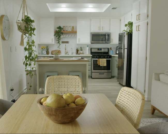 A kitchen and dining area feature a wooden table with a fruit bowl, surrounded by wicker chairs, while the kitchen has white cabinets and stainless steel appliances.