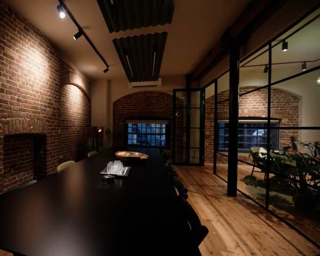 The image shows a modern dining area with a long black table, exposed brick walls, and wooden flooring, illuminated by track lighting.