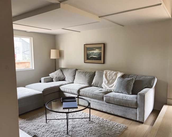 A light-filled living room features a gray sectional sofa, a circular glass coffee table, and a framed landscape painting on the wall.