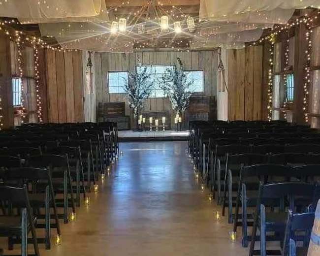 The interior of a rustic venue is set up for a wedding ceremony, featuring rows of black chairs, decorative lighting, and a decorated altar at the far end.