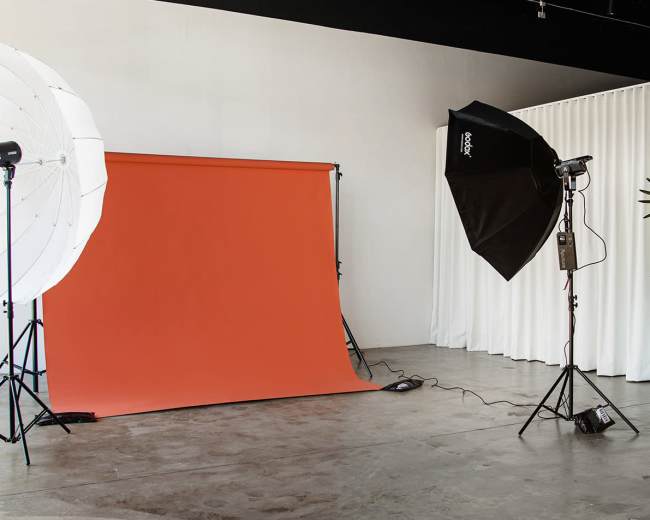 The image shows a photography studio setup with an orange backdrop, two light sources, and a potted plant in the corner.