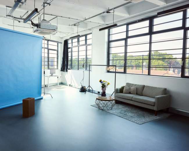 The image shows a bright studio space featuring a blue backdrop, a sofa, a coffee table, and large windows allowing natural light to enter.