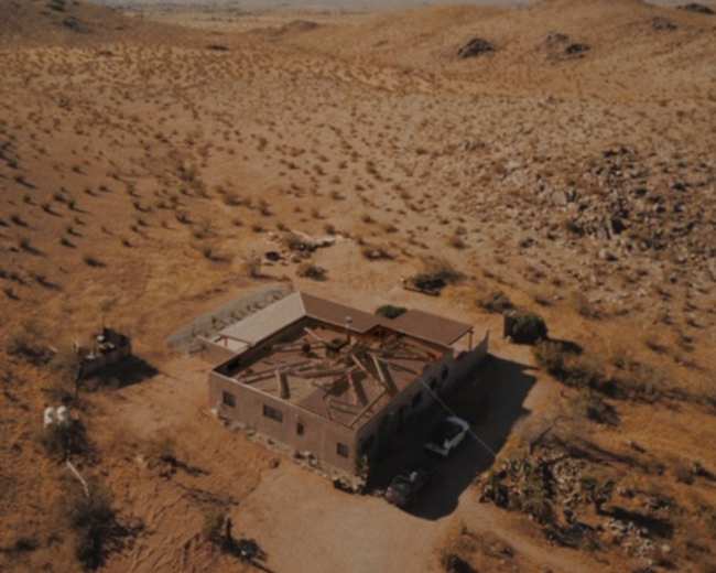 An abandoned building sits in a vast desert landscape, surrounded by rolling hills and sparse vegetation.