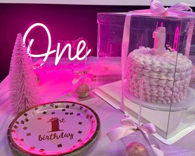 A decorated birthday table features a pink "One" neon sign, a clear cake box with a one-shaped cake, themed plates, and cupcakes.