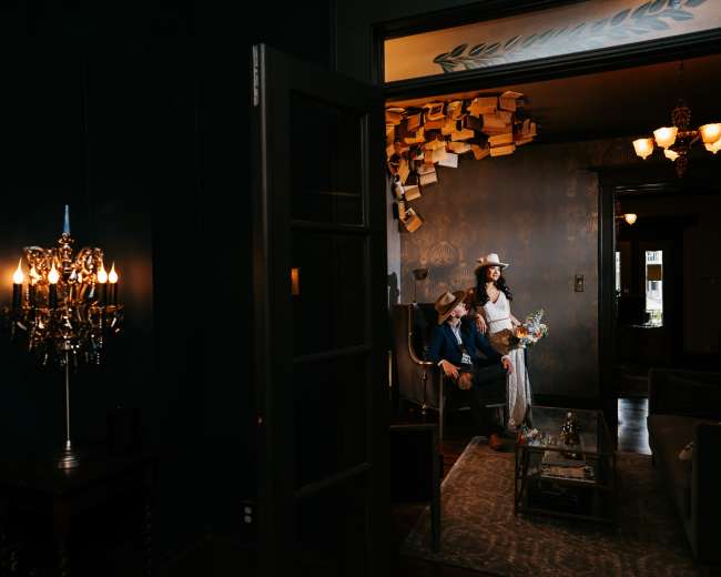 A couple dressed in formal attire is seated together in a dimly lit room with books arranged artistically on the walls.