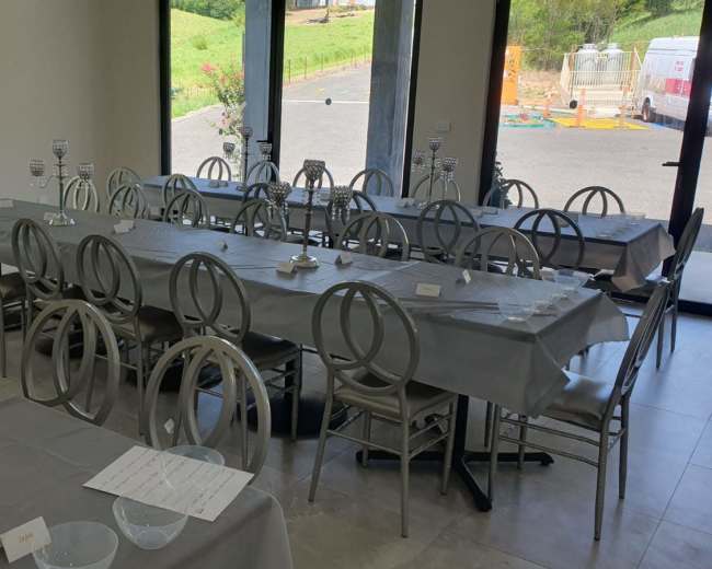 A large room features several long tables covered with gray tablecloths and arranged with silver chairs, set for an event with candelabras and place cards.