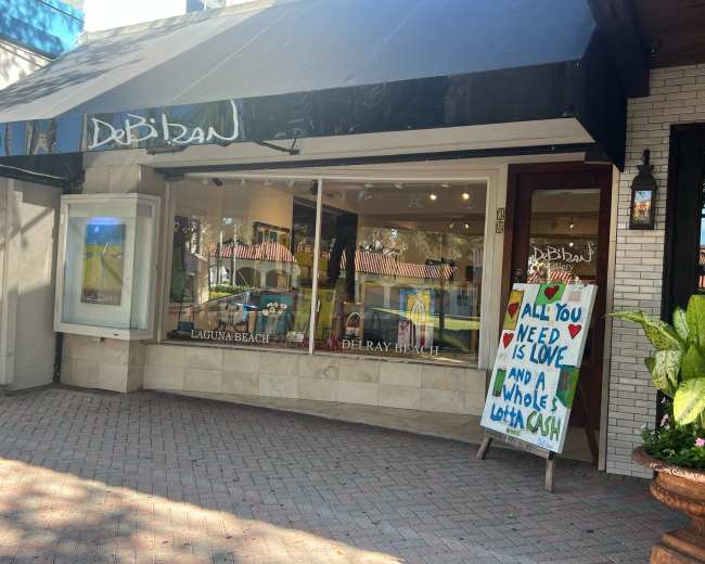 The image shows a storefront with large windows displaying colorful art and a sidewalk sign reading "All You Need is Love and a Whole Lotta Cuch."