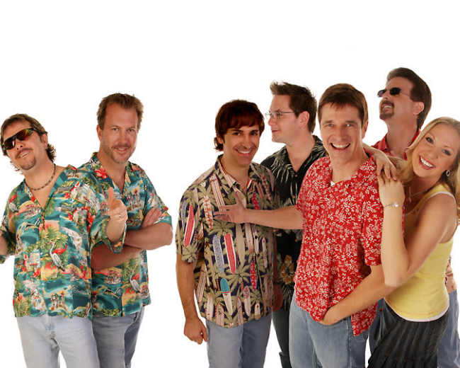 A group of six men and one woman stands together, wearing colorful tropical shirts against a white background.