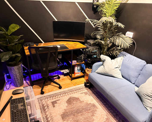 The image shows a cozy office space featuring a wooden desk with a computer monitor, a black office chair, a blue sofa, a potted plant, and various storage items.