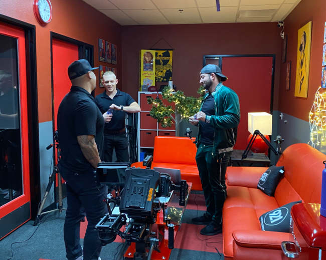 Three men are talking in a room with red walls and a red sofa, surrounded by camera equipment and decorations.