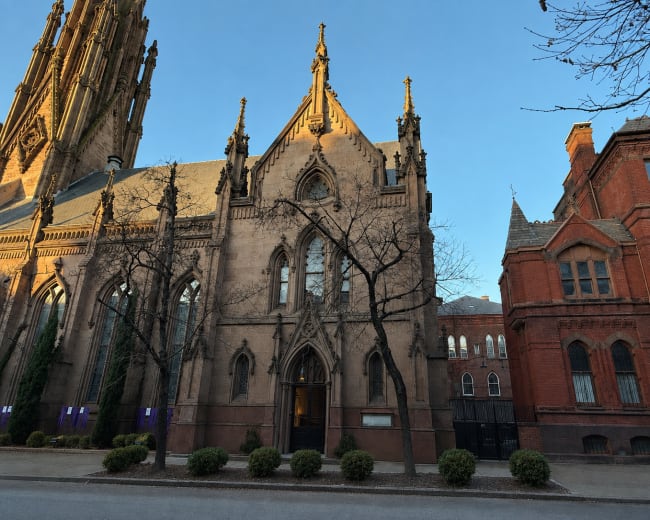 The image shows a tall Gothic church with intricate architectural details alongside a brick building, set against a clear sky.