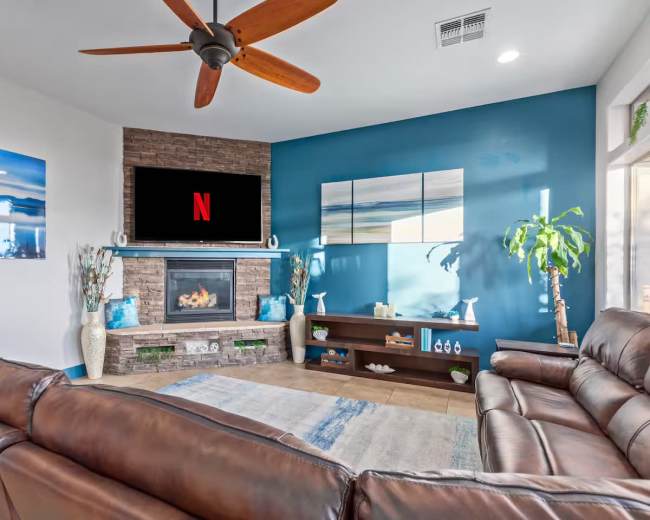 The image shows a modern living room with a leather sectional, a stone fireplace, a mounted TV displaying the Netflix logo, and decorative plants against a blue accent wall.