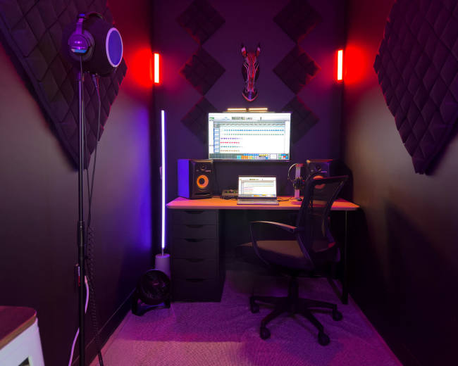The image shows a small, dark recording studio featuring a desk with a computer, speakers, and a headset, illuminated by colorful LED lights and decorated with sound-absorbing panels.