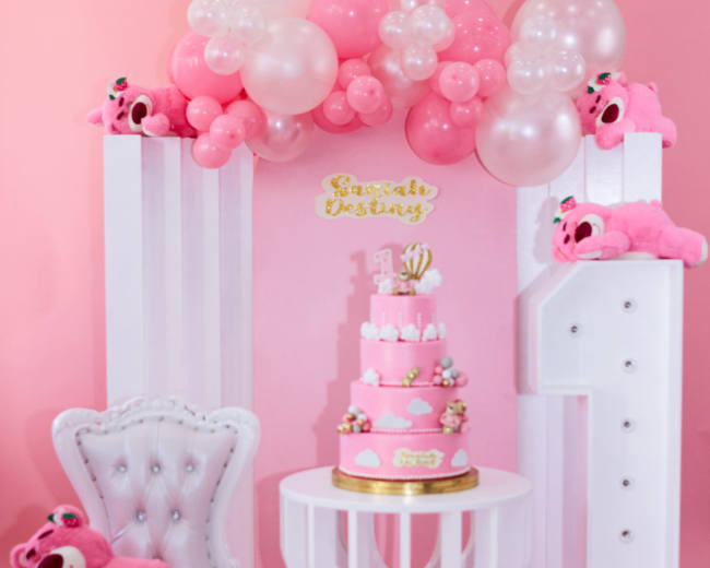 A pink-themed birthday setup features a large cake on a table surrounded by plush toys and balloon decorations.