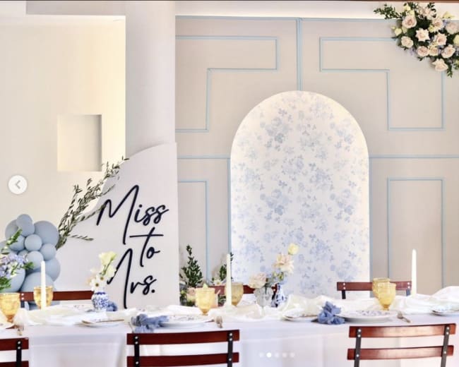 A decorated dining table is set against a white wall featuring floral patterns and an arch, with a sign that reads "Miss to Mrs." on the side.