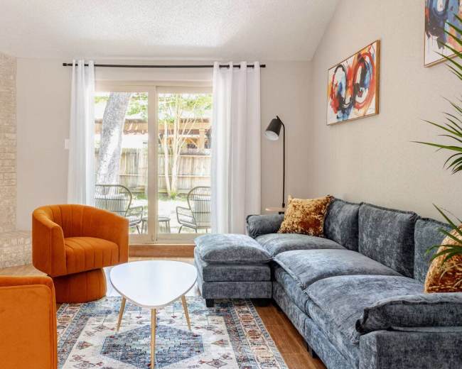 A modern living room features a blue sectional sofa, orange accent chairs, a round coffee table, and large windows overlooking a backyard.