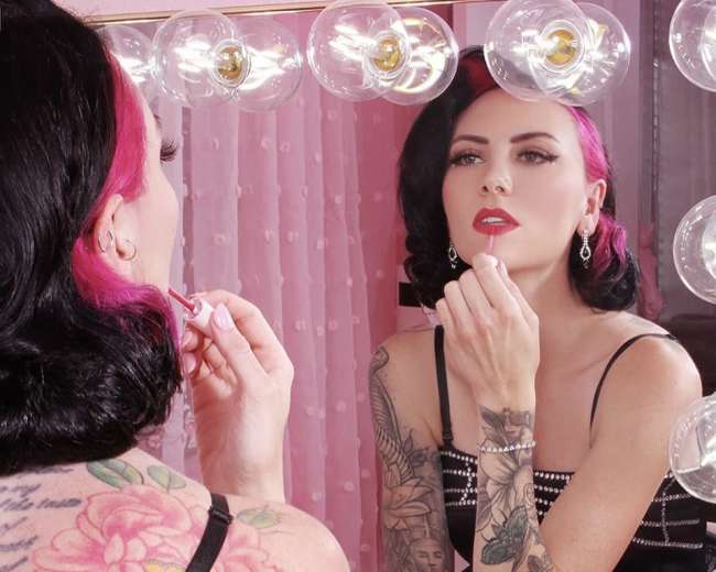 A woman with pink highlights applies lipstick while sitting in front of a mirror surrounded by decorative figurines and illuminated vanity lights.