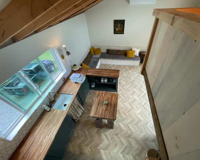 The image shows an open-concept kitchen and living area with wooden countertops, a herringbone-patterned floor, and a large window allowing natural light to enter.