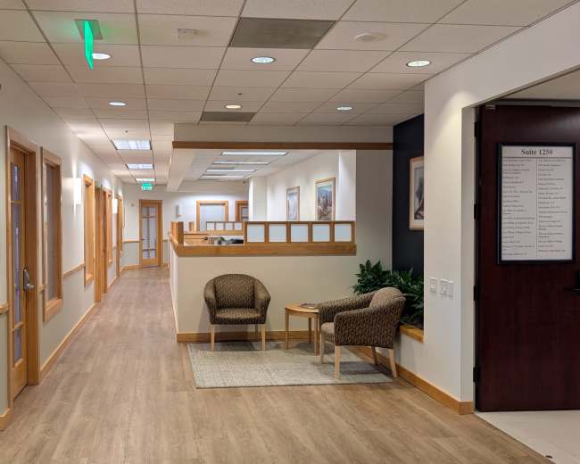 The image shows a well-lit hallway with wooden accents, featuring two chairs and a small table near the entrance to various office suites.