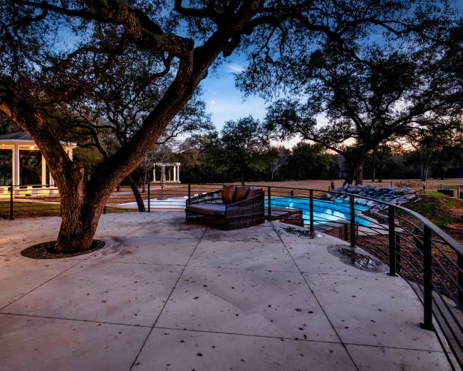 The image shows a patio area featuring a large tree, a seating arrangement, and a swimming pool surrounded by landscaped greenery.