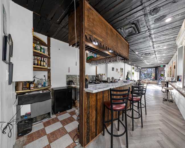 The image shows a dimly lit cafe or bar interior featuring a wooden counter with high stools, shelves stocked with bottles, and a tiled floor.