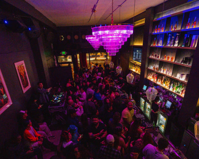 A large crowd mingles inside a dimly lit bar featuring a vibrant chandelier and shelves filled with colorful bottles.
