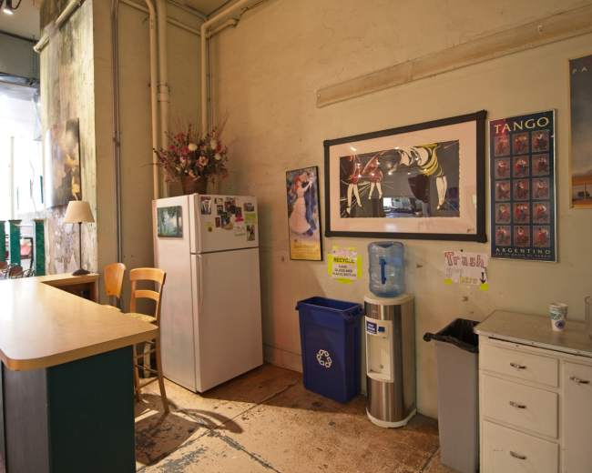 The image shows a kitchen area with a refrigerator, water cooler, recycling bin, and various posters on the walls.