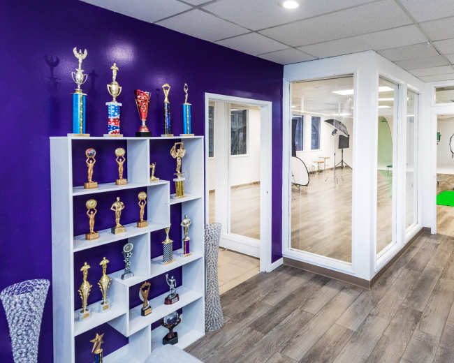 A brightly lit interior space with purple walls, featuring a trophy display on the left and a large glass-walled room in the background.