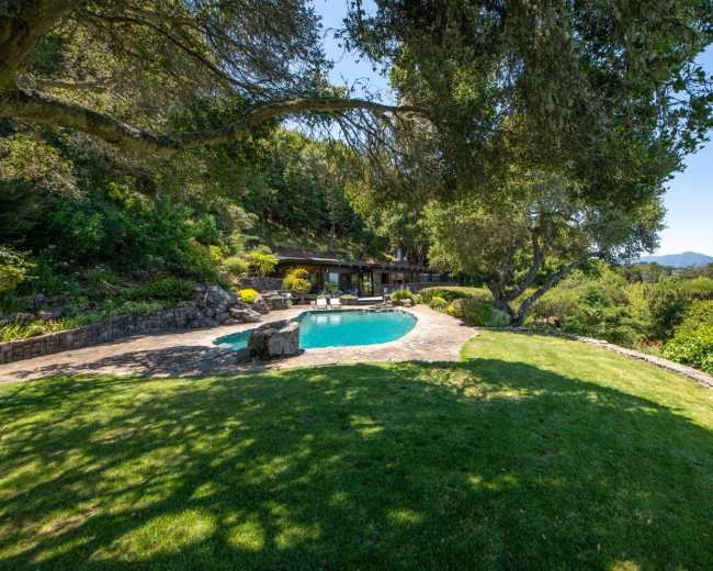 A swimming pool is surrounded by lush greenery and stone pathways, with a house nestled in the hillside in the background.